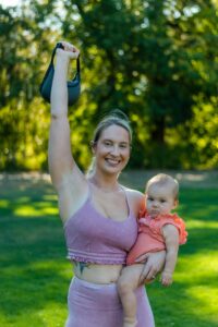 Smiling woman exercising outdoors with baby, holding a kettlebell.